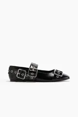 Zapatos planos tipo bailarina o mary jane, color negro charol, con punta redonda. Presentan una tira sobre el empeine adornada con múltiples ojales metálicos y una hebilla grande de metal.