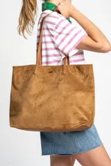 Bolso tipo shopper de gran tamaño confeccionado en gamuza vegana color camel. Presenta dos asas largas para el hombro, base reforzada y una pequeña placa metálica frontal con el logo de la marca.