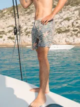 Short de baño con estampado botánico de hojas en tonos verdes y salmón sobre fondo gris azulado. Cuenta con cintura elástica con cordón ajustable, dos bolsillos laterales y un bolsillo trasero.