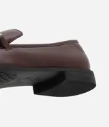 Mocasines de mujer confeccionados en poliuretano color marrón oscuro, con acabado brillante y detalle decorativo metálico dorado en el empeine. Presentan una silueta clásica de punta redondeada y suela plana con pequeño tacón.