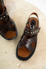 Sandalias de cuero marrón con textura de cocodrilo, diseño de tiras cruzadas con tachas metálicas plateadas y suela track negra de plataforma.