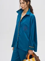 Camisa azul de corte holgado, con cuello camisero, mangas largas con detalles en los puños, bolsillo frontal con bordado y cierre delantero con botones.