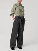 Blusa verde de algodón orgánico con cuello camisero, manga larga y cierre frontal de botones. Presenta volantes bordados que recorren el frente, la espalda y las sisas, además de detalles bordados en toda la prenda.