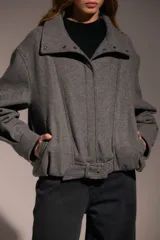 Campera tipo bomber de corte holgado y diseño estructurado, confeccionada en tejido gris con textura de espiga. Presenta cuello alto con cierre de broches a presión, mangas largas con puños ajustados y un cinturón integrado en el ruedo con hebilla metálica para un ajuste personalizado.