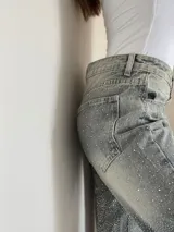 Pantalón de jean rígido de corte recto, con acabado lavado y aplicación de brillos distribuidos por toda la prenda. Presenta diseño de cinco bolsillos, cierre frontal con botón y tiro medio.