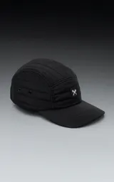 Gorra negra de estilo 5 paneles, con tejido acolchado o con costuras horizontales y visera plana. Presenta un pequeño parche cuadrado negro con un logo blanco bordado en forma de 'X' o lazo.