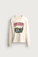 Camiseta de manga larga y cuello redondo en color crudo, con estampado gráfico frontal de paisaje montañoso y tipografía en tonos granate y verde.