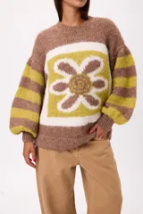Buzo de tejido de punto con textura peluda, de corte holgado y mangas amplias. Presenta un diseño geométrico con una flor grande estampada en el centro, combinando tonos fucsia, rosa y marrón en un patrón de bloques y rayas.