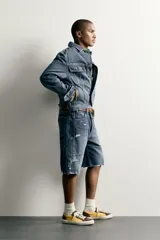 Bermuda baggy fit confeccionada en denim de algodón con lavado azul y efecto rotos en toda la prenda. Presenta diseño de cinco bolsillos, detalles de costuras en las perneras, bajo acabado irregular y cierre frontal con cremallera y botón.