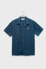Camisa azul con bordado de flores en el pecho, de corte cuadrado y calce relajado. Confeccionada en Italia en mezcla de lino y algodón. Tiene cuello tipo camp y cierre con botones de coco grabados con el logo.