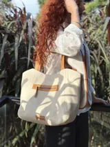Bolso tipo tote color crema, con detalles en cuero color marrón. Tiene un bolsillo con cierre en la parte superior y un parche de cuero con el logo de la marca en la parte inferior.