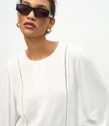 Blusa tipo bata de color blanco, confeccionada en viscosa, con cuello redondo, mangas largas con puños abotonados, detalles de entremedios verticales y un recorte con volado en el bajo.