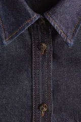 Camisa de denim de algodón rígido en color azul oscuro, con cuello clásico, cierre frontal de botones, bolsillos de parche en el pecho, mangas largas con puños abotonados y bajo redondeado.