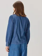 Camisa de denim con silueta levemente entallada, cuello bebé y mangas largas con sutil frunce en los hombros. Presenta costuras curvas decorativas con pespuntes en contraste y cierre frontal con botones.