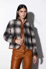 Chaqueta corta de paño a cuadros en tonos beige y negro, con cuello mao y ribete de cuero marrón en todo el contorno. Se ata con lazos de cuero en el centro y tiene bolsillos laterales.
