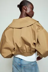 Chaqueta corta de Zara confeccionada en mezcla de algodón, con cuello solapa, manga larga con hombro caído y puños elásticos. Presenta un diseño cruzado con cierre de hebilla metálica lateral y forro interior a tono.