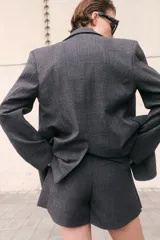 Blazer de mujer gris oscuro con estampado Príncipe de Gales (a cuadros). Presenta solapas de muesca, cierre frontal de dos botones, hombreras y bolsillos delanteros con solapa.