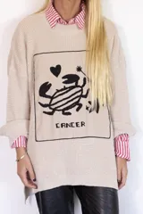 Sweater de punto oversize color crudo, con cuello redondo y diseño bordado a mano en el frente representando al signo Cáncer con un cangrejo, un corazón y una estrella.
