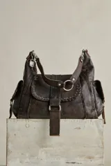 Cartera de cuero de formato hobo, con efecto gastado, correa de hombro ajustable, solapa frontal con argolla metálica y detalles de costuras y ojales metálicos.