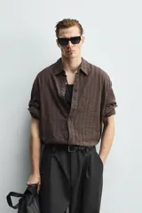 Camisa de corte relajado confeccionada en tejido ligero de algodón con textura de cuadros. Presenta cuello solapa, manga larga con puños abotonados y cierre frontal de botones.
