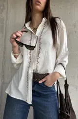 Camisa blanca de tejido liviano y calce relajado, con manga larga y detalle de puntilla y bordado en contraste a lo largo de la botonadura frontal.