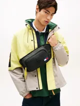 Ri±onera negra Tommy Jeans de diseño urbano, confeccionada en poliuretano y poliéster. Cuenta con un compartimento principal, bolsillo exterior con cierre de cremallera, tiradores en color amarillo, asa superior, correa ajustable y el parche bordado con el logotipo de la marca.