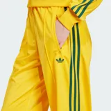 Pantalón deportivo Adidas de corte holgado, color amarillo brillante con las clásicas tres tiras verdes en los laterales y el logo del trifolio bordado en la pierna izquierda. Cuenta con cintura elástica y bolsillos laterales con cierre.