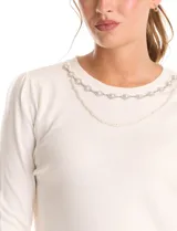 Buzo de tejido fino color blanco, con escote redondo decorado con un collar fijo de perlas y strass. Presenta mangas largas con un leve frunce en los hombros y puños acanalados.