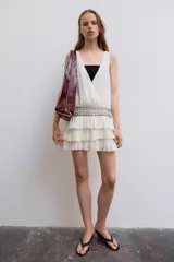 Vestido corto de gasa color blanco, con escote en V cruzado, sin mangas y diseño con volantes en el bajo. Presenta un detalle de tachas plateadas en la cintura.