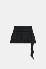 Falda pantalón de tiro bajo en color negro, con cintura drapeada y detalle de tira lateral colgante. Presenta un diseño con volantes en el bajo y cierre de cremallera metálica.
