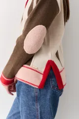 Buzo de punto con cuello polo y cierre frontal de tres botones. Presenta un diseño a rayas verticales en tonos beige y marrón, con mangas en bloques de color rojo y detalles en rosa en los puños y el ruedo.