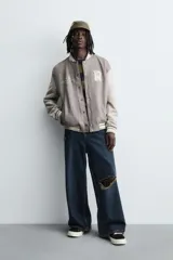 Cazadora estilo varsity o bomber de hombre, color gris oscuro con efecto ante, mangas en tono gris claro y puños, cuello y bajo con rib elástico a rayas blancas y grises. Presenta un parche con la letra 'R' en el pecho derecho y un bordado con texto en el pecho izquierdo.