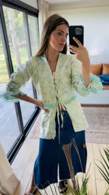 Camisa de brocato verde clara con estampado floral, escote en V y cierre frontal con lazos ajustables. Presenta mangas tres cuartos con detalles de fruncido tipo smock y terminaciones con flecos en los puños y el borde inferior.