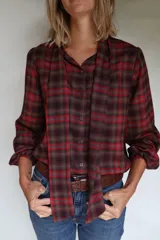 Camisa de estilo romántico con estampado escocés en tonos rojos y marrones. Presenta cuello a la base con lazos integrados, mangas largas con frunce en los hombros y puños elásticos. Incluye cierre frontal con botones de coco.