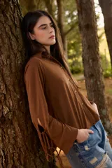 Camisa de color marrón camel con cuello clásico y cierre frontal de botones. Presenta mangas largas con aberturas laterales y detalles de tiras para atar en los puños.