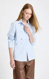 Camisa de algodón color celeste con cuello clásico, manga larga con puños y cierre frontal de botones. Presenta apliques decorativos de pedrería brillante distribuidos por toda la prenda.