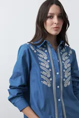 Camisa de manga larga en tono azul denim, con cuello clásico y cierre frontal de botones. Presenta un delicado bordado floral en color crema que enmarca la botonadura central, aportando un estilo artesanal y sofisticado.