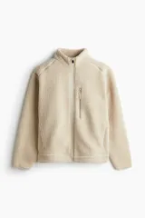 Campera deportiva de tejido sherpa color beige, con cuello elevado, cierre frontal completo, mangas raglán, bolsillo superior con cierre y bolsillos laterales. Presenta terminaciones elásticas en puños y ruedo.