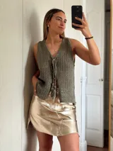 Chaleco tejido en punto calado con hilo de algodón y lurex, de color negro con destellos plateados. Presenta escote profundo en V, cierre frontal con cordones entrelazados terminados en borlas y un corte recto y holgado.