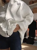 Campera tipo hoodie oversize de textura tipo corderito en color blanco, con diseño de parches en la espalda que combinan tela de jean a rayas y encaje blanco. Cuenta con capucha y un pequeño parche de cuero sintético en la parte inferior trasera.