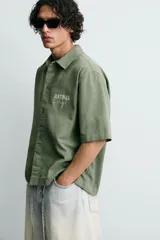 Camisa de corte relajado color verde oliva, con cuello solapa, manga corta y cierre frontal de botones. Presenta un estampado con el texto 'ASTRAL Drift' en el frente.