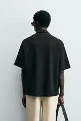 Camisa de corte relajado en color negro, confeccionada en tejido de algodón. Presenta cuello tipo bowling, manga corta y cierre frontal con botones. Cuenta con bordados lineales de hojas en la parte delantera.