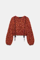 Jersey de punto grueso con textura trenzada en color naranja y bordó. Presenta cuello redondo, mangas largas abullonadas y detalle de cordones trenzados a contraste en el frente con terminación en lazada en el ruedo.