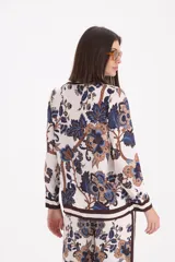 Camisa de manga larga con cuello clásico y cierre frontal de botones. Presenta un estampado floral en tonos beige, marrón y azul, con detalles de bordes en color marrón oscuro.