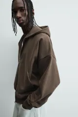 Sudadera con capucha y cierre de cremallera frontal, color marrón oscuro, de corte holgado (boxy fit) y manga larga. Presenta bolsillos frontales de vivo y acabados en rib en puños y bajo.