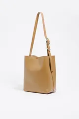 Bolso de hombro tipo cubo de estructura rígida, color marrón oscuro, confeccionado en piel vacuna. Posee asa regulable con hebilla metálica y cierre de cordón en la parte superior. Lleva el logo metálico de Bimba y Lola en la parte frontal inferior.