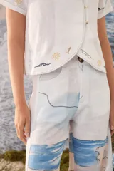 Blusa blanca de lino con mangas cortas, cuello alto con volado y cierre lateral cruzado con botones dorados. Presenta detalles bordados artesanales a contratono en el ruedo y las mangas.