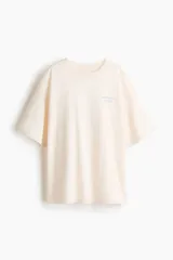 Remera oversized de punto de algodón color beige claro, con cuello redondo acanalado, hombros caídos y moldería amplia. Presenta un pequeño estampado en el pecho con el texto "Taormina Sicily" en color celeste.