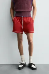 Short de algodón color rojo oscuro, de corte regular fit, con cintura ajustable mediante cordón blanco. Presenta ribetes o vivos de contraste en color blanco bordeando los laterales, el bajo y los bolsillos.