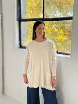 Sweater de punto color beige, de corte holgado y diseño oversize. Presenta cuello redondo, mangas largas con puños ajustados y dos bolsillos frontales tipo parche. El diseño incluye una costura vertical central en el frente y terminaciones acanaladas en el ruedo.